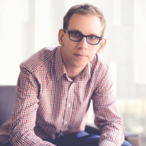 Michael Kutcher, white male, blonde with glasses is sitting and leaning slightly front and wearing a lilac button-down shirt.