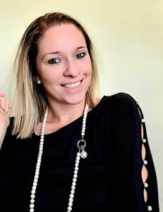 Blonde woman with brown eyes who is smiling, wearing a black shirt and pearl necklace.